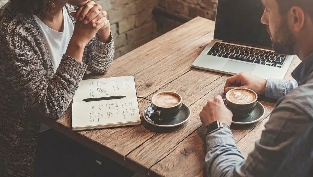 A laptop, open notebook, and two coffees on a wooden table during a work meeting